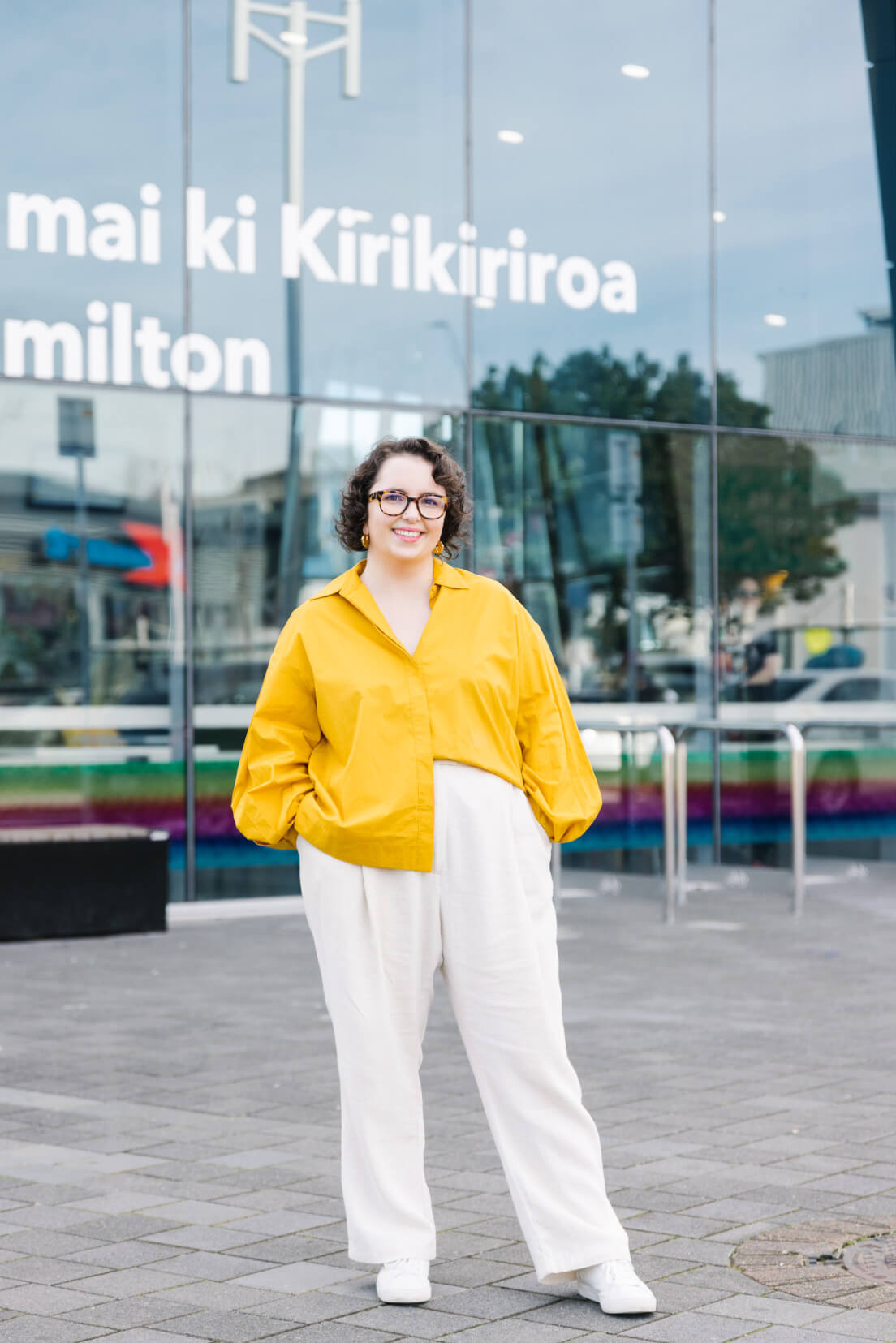 Louise, wearing a bright yellow shirt, standing in front of the glass walls of the Hamilton Transport Centre